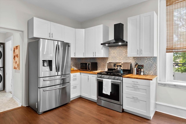 kitchen featuring wall chimney range hood, appliances with stainless steel finishes, stacked washing maching and dryer, dark wood-type flooring, and butcher block counters
