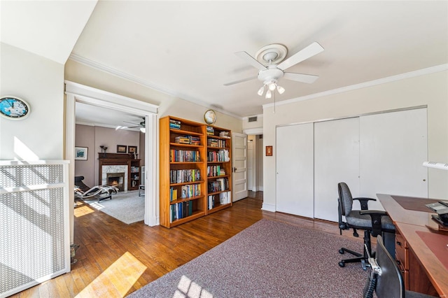 office featuring a warm lit fireplace, visible vents, hardwood / wood-style flooring, ceiling fan, and crown molding