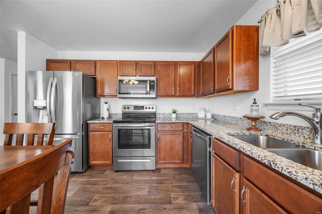 kitchen with brown cabinetry, dark wood finished floors, light stone counters, appliances with stainless steel finishes, and a sink