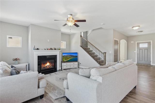 living area with stairs, ceiling fan, a glass covered fireplace, and wood finished floors