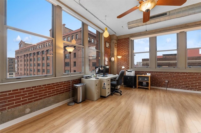 office area with light wood-type flooring, a healthy amount of sunlight, visible vents, and brick wall