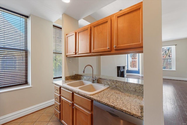 kitchen featuring light stone counters, brown cabinetry, a sink, dishwasher, and baseboards