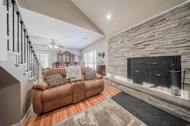 living area featuring wood finished floors, lofted ceiling, a stone fireplace, stairway, and a ceiling fan