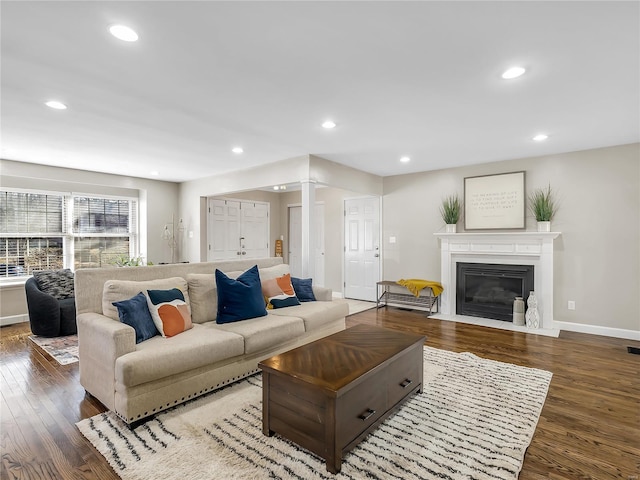 living room featuring wood finished floors, baseboards, a fireplace with flush hearth, recessed lighting, and decorative columns