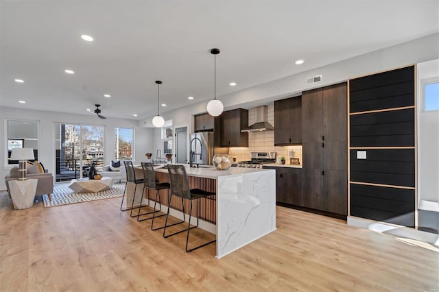 kitchen with pendant lighting, stove, wall chimney range hood, dark brown cabinets, and an island with sink