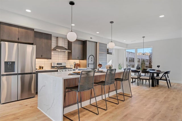 kitchen featuring a breakfast bar, appliances with stainless steel finishes, wall chimney exhaust hood, light wood finished floors, and a center island with sink