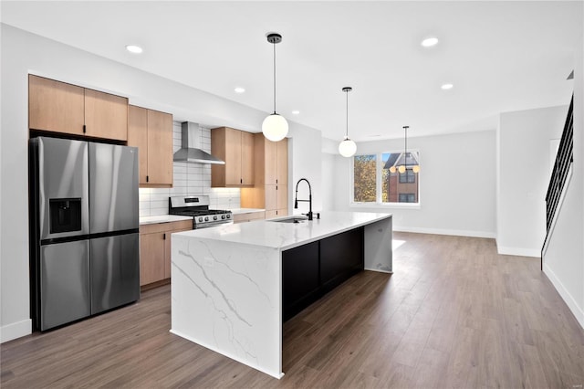 kitchen with a center island with sink, hanging light fixtures, sink, appliances with stainless steel finishes, and wall chimney exhaust hood