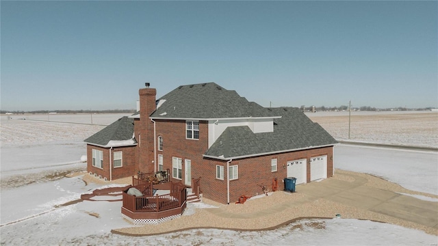 snow covered property with driveway, brick siding, a chimney, and a shingled roof