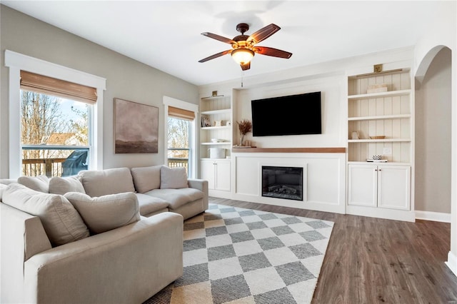 living area with ceiling fan, arched walkways, built in shelves, a glass covered fireplace, and dark wood finished floors