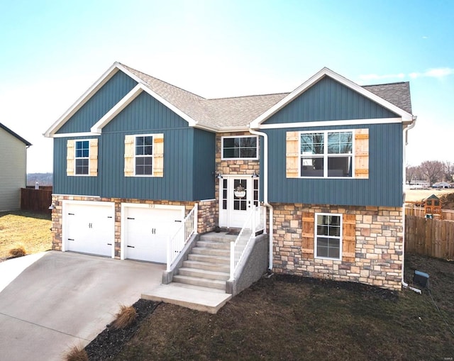 bi-level home featuring driveway, a garage, stone siding, roof with shingles, and fence