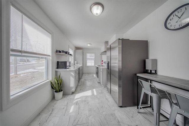 kitchen with a sink, marble finish floor, appliances with stainless steel finishes, wall chimney range hood, and light countertops