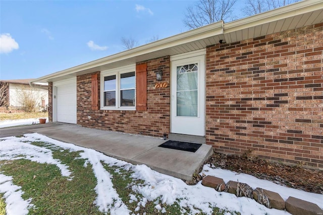 snow covered property entrance featuring brick siding, driveway, and an attached garage