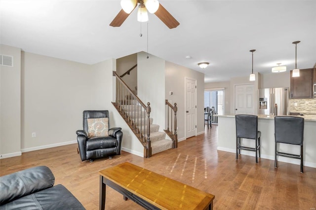 living area featuring light wood-type flooring, visible vents, baseboards, ceiling fan, and stairs