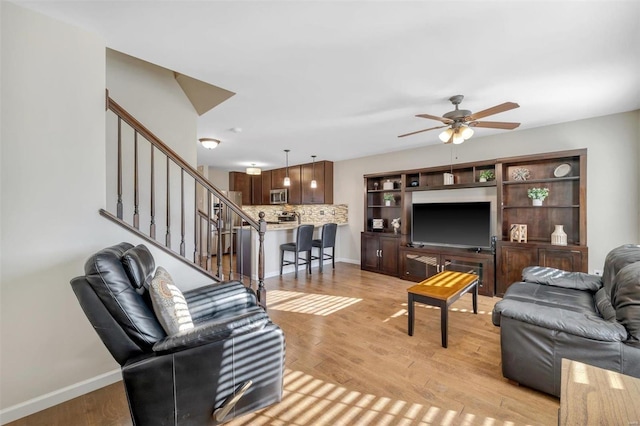 living room featuring stairs, baseboards, light wood-style floors, and a ceiling fan