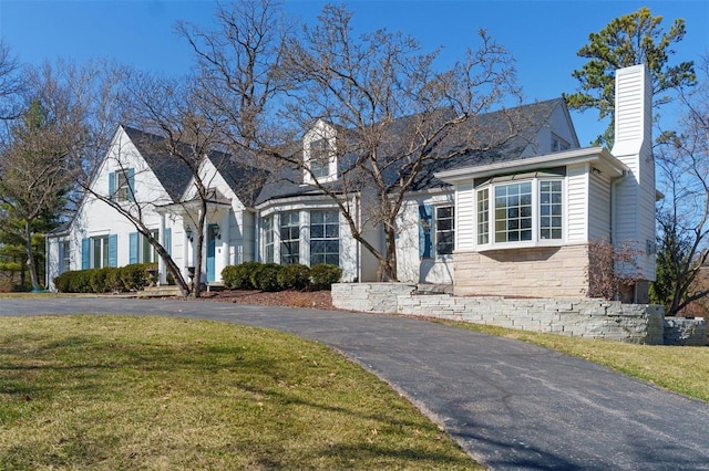 cape cod-style house featuring driveway, stone siding, a chimney, and a front yard