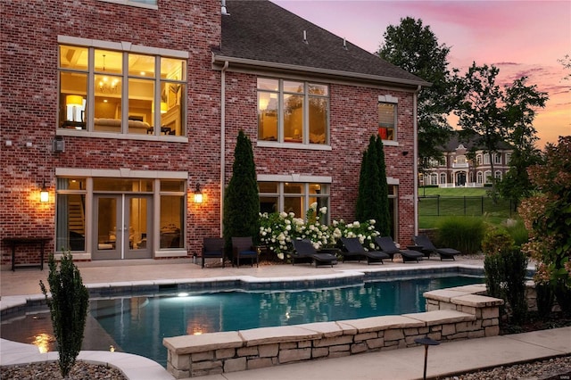 back of house at dusk featuring an outdoor pool, a patio area, fence, and brick siding