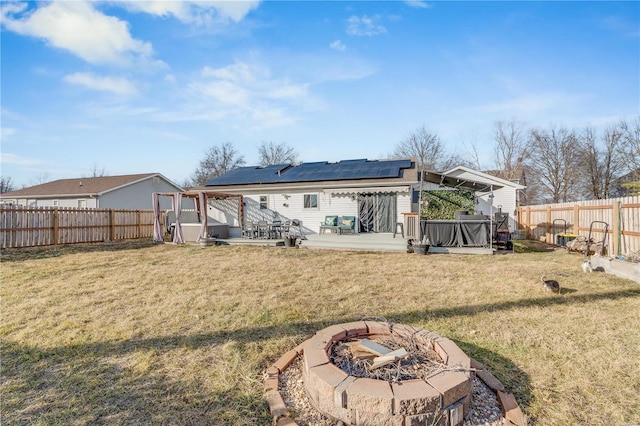 rear view of property with a deck, a fenced backyard, a fire pit, a lawn, and roof mounted solar panels