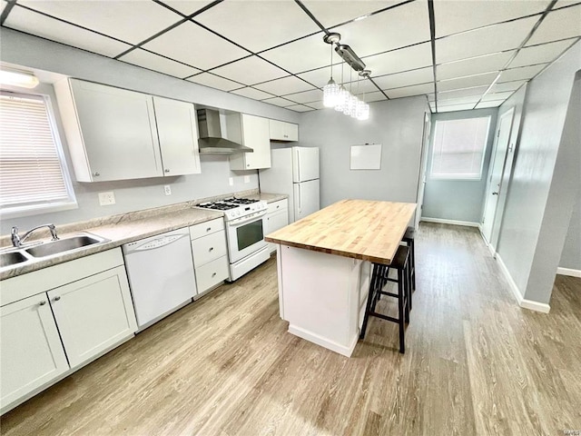 kitchen with white appliances, a sink, white cabinets, hanging light fixtures, and wall chimney range hood