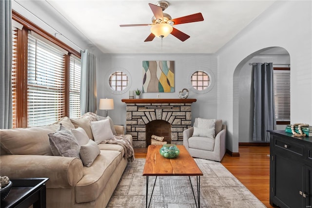 living room featuring arched walkways, light wood-style floors, a ceiling fan, a stone fireplace, and baseboards