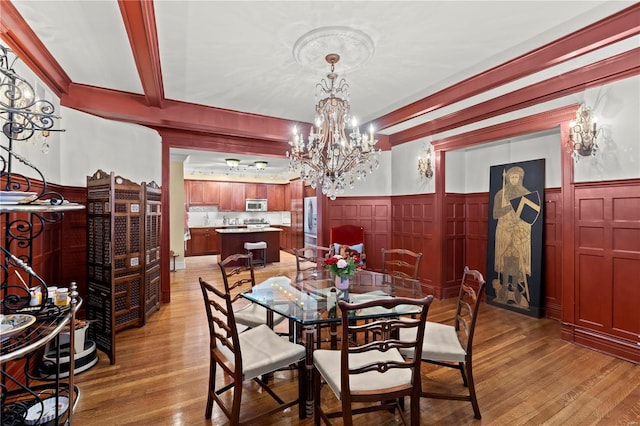 dining space with light wood-type flooring, an inviting chandelier, beamed ceiling, and wainscoting