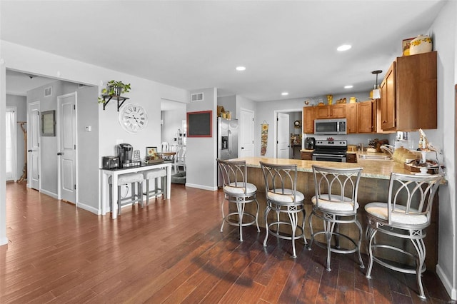 kitchen featuring dark wood finished floors, appliances with stainless steel finishes, brown cabinets, decorative light fixtures, and a peninsula