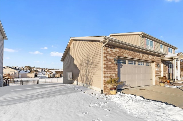 snow covered property featuring a garage, brick siding, fence, and driveway