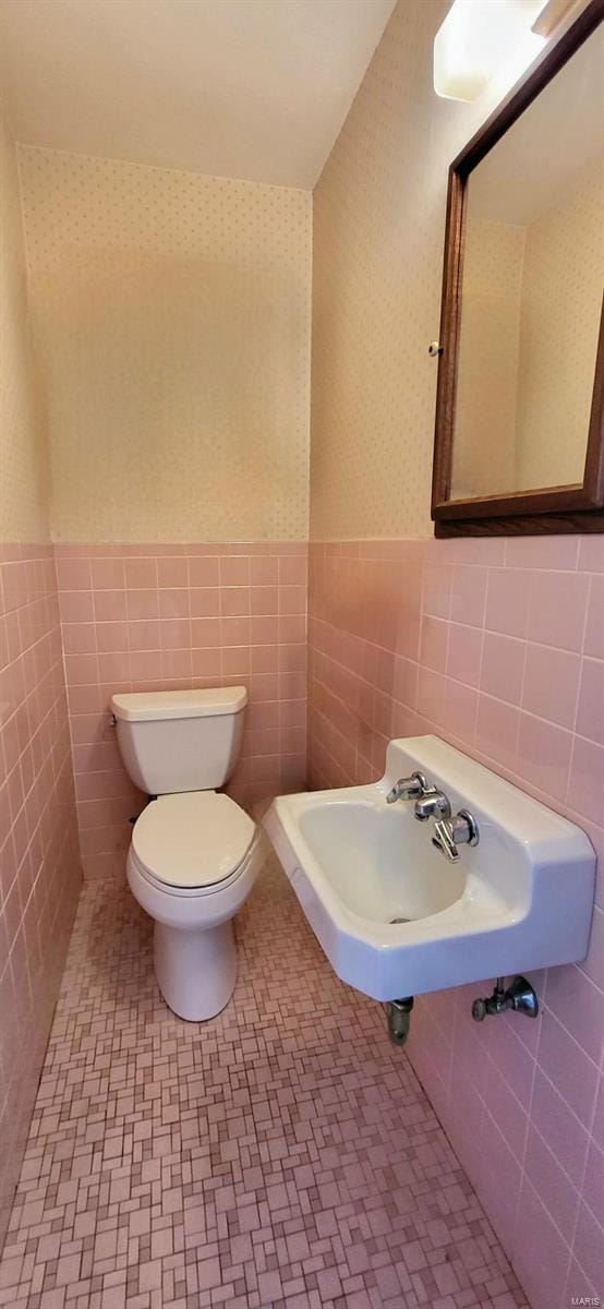 bathroom featuring wallpapered walls, wainscoting, toilet, a sink, and tile walls