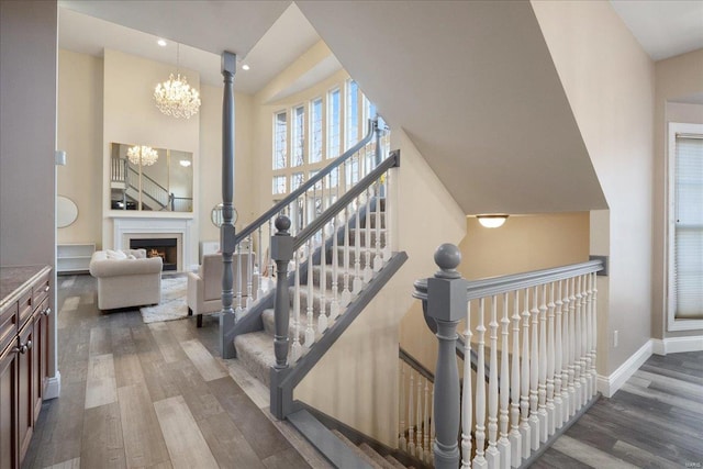 staircase with a notable chandelier, a towering ceiling, wood finished floors, a lit fireplace, and baseboards