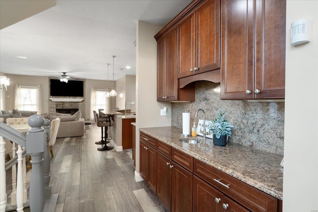 kitchen with ceiling fan with notable chandelier, a fireplace, wood finished floors, a sink, and light stone countertops