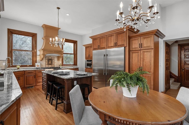 kitchen featuring a chandelier, stainless steel appliances, brown cabinets, plenty of natural light, and custom range hood