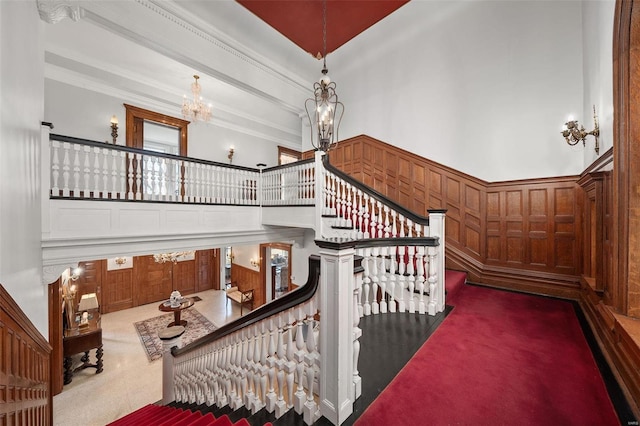 staircase featuring carpet floors, crown molding, a decorative wall, a high ceiling, and a chandelier