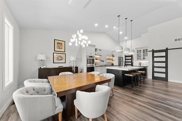 dining area featuring visible vents, a barn door, wood finished floors, high vaulted ceiling, and baseboards