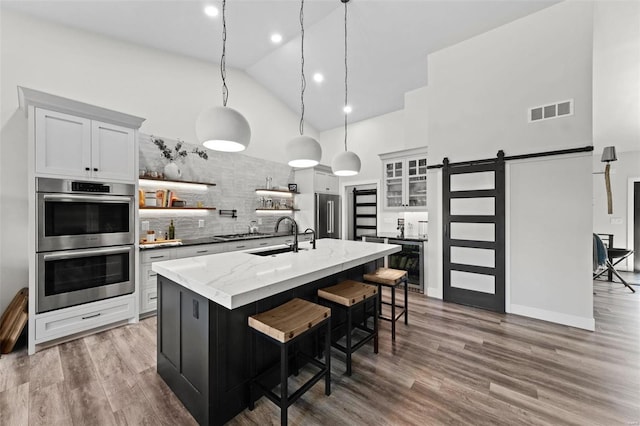 kitchen with a barn door, visible vents, glass insert cabinets, a kitchen island with sink, and stainless steel appliances