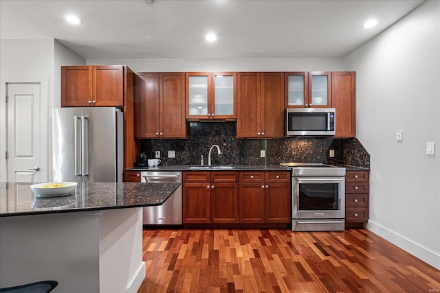 kitchen with dark wood-style flooring, a sink, appliances with stainless steel finishes, dark stone counters, and glass insert cabinets