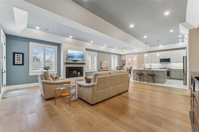 living area featuring light wood-style flooring, a warm lit fireplace, a raised ceiling, and baseboards