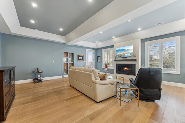 living room featuring visible vents, baseboards, a tray ceiling, a warm lit fireplace, and light wood-style floors