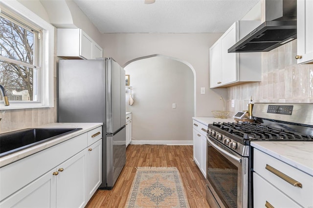 kitchen with arched walkways, stainless steel appliances, white cabinets, a sink, and wall chimney range hood