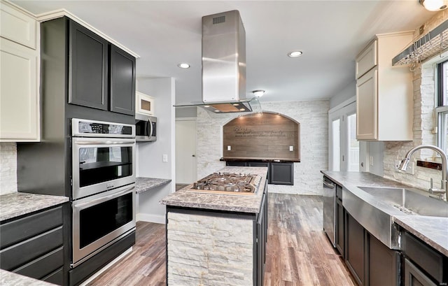 kitchen with backsplash, appliances with stainless steel finishes, a sink, island range hood, and wood finished floors