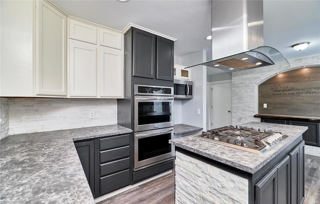 kitchen with island range hood, stainless steel appliances, wood finished floors, a kitchen island, and decorative backsplash