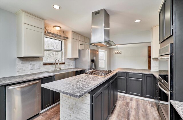 kitchen with island exhaust hood, stainless steel appliances, decorative backsplash, light wood-style floors, and a sink