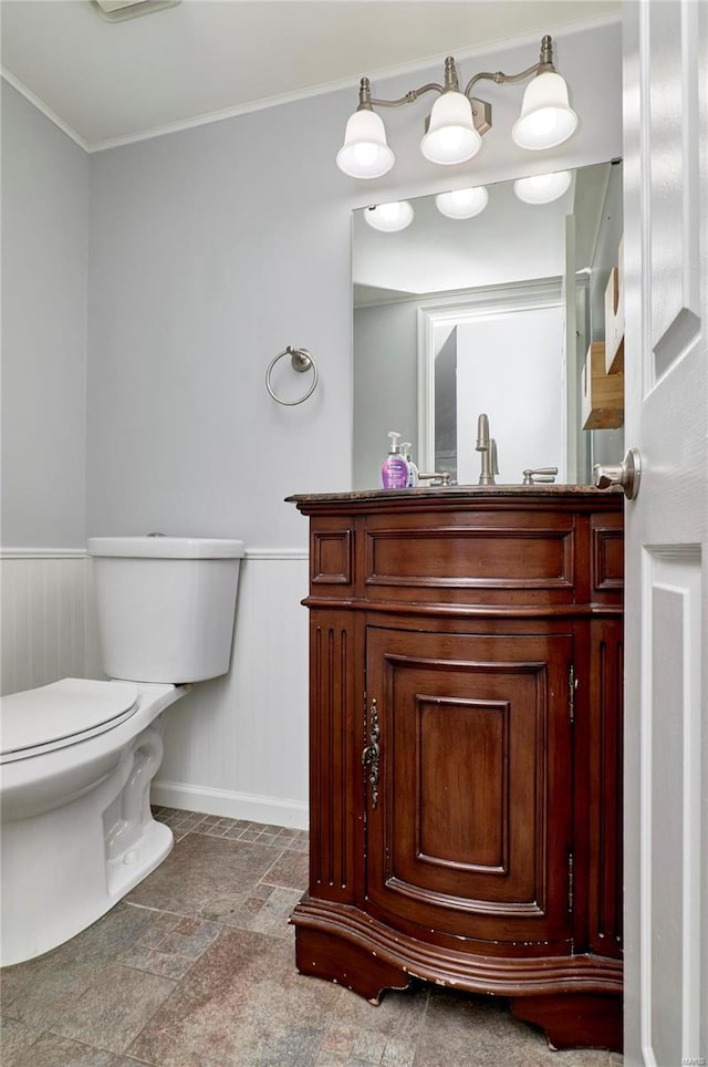 half bath featuring toilet, ornamental molding, stone finish flooring, wainscoting, and vanity