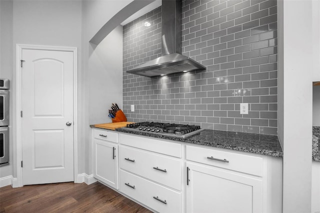 kitchen with dark stone countertops, dark wood-style floors, appliances with stainless steel finishes, wall chimney exhaust hood, and backsplash