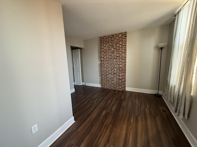 empty room with brick wall, dark wood-style flooring, visible vents, and baseboards