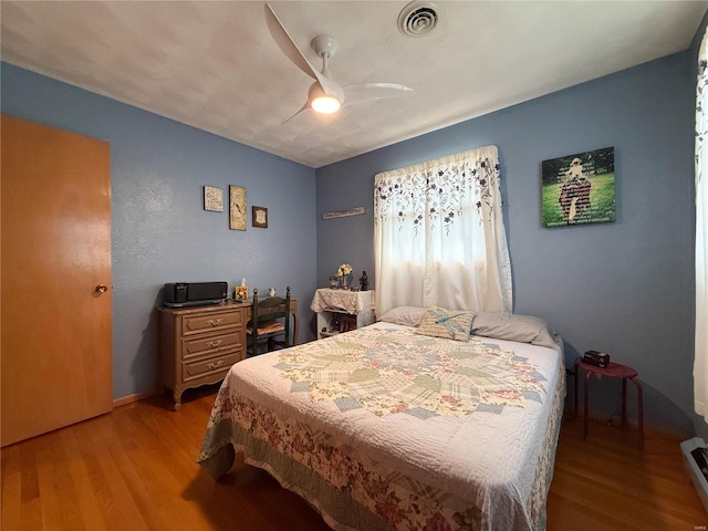 bedroom featuring baseboard heating, wood finished floors, visible vents, and a ceiling fan