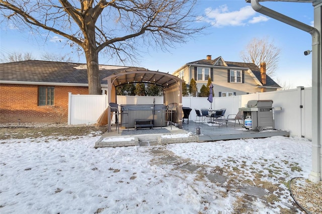 snow covered back of property with a deck, brick siding, a fenced backyard, and a hot tub