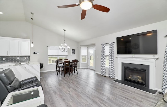 living area with a glass covered fireplace, vaulted ceiling, light wood-type flooring, baseboards, and ceiling fan with notable chandelier