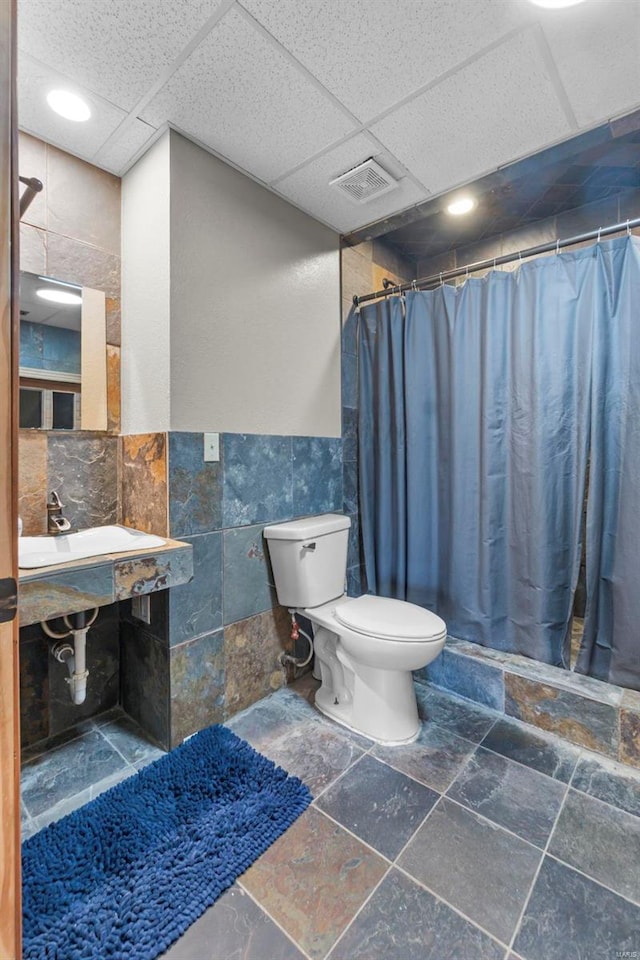 bathroom with a drop ceiling, toilet, stone tile floors, visible vents, and tile walls