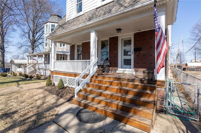 property entrance featuring fence, brick siding, covered porch, and roof with shingles