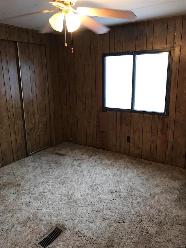 carpeted empty room featuring a ceiling fan, visible vents, and wooden walls