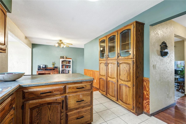 kitchen featuring light tile patterned flooring, brown cabinetry, a ceiling fan, and wainscoting